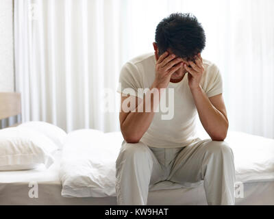 asian man painful and miserable sitting on bed head down covering face with hands. Stock Photo