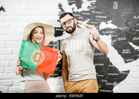 Couple of young travelers standing with portuguese flag near the wall with world map, dreaming about summer vacations in Portugal Stock Photo