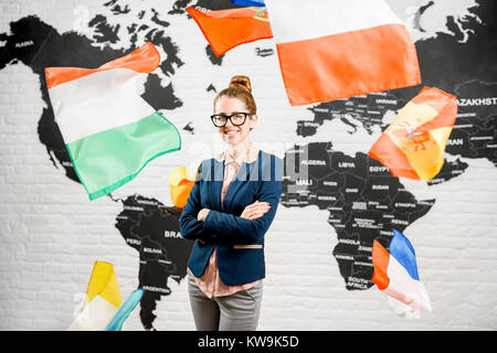Portrait of a female travel agent dressed in the suit standing indoors surrounded by flying flags on the world map background Stock Photo