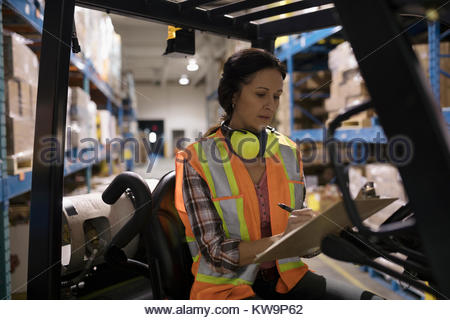 Female worker with clipboard driving forklift in distribution warehouse