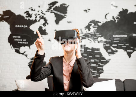 Young businesswoman trying virtual reality glasses sitting indoors with world map on the background Stock Photo