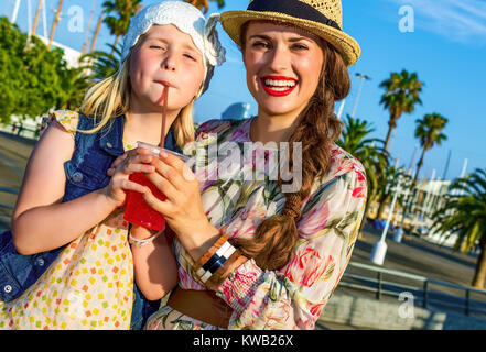 Summertime at colorful Barcelona. smiling elegant mother and daughter tourists on embankment in Barcelona, Spain drinking bright red beverage Stock Photo