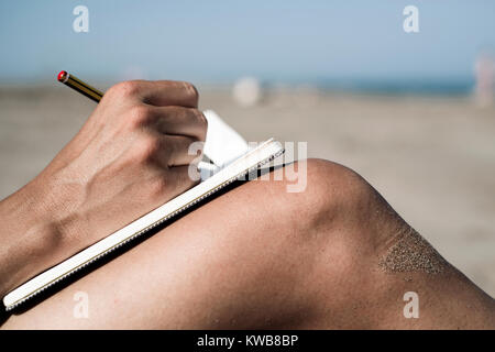 closeup of a young caucasian man writing with a pen in a notebook on his shaved legs, on the beach during the summer Stock Photo