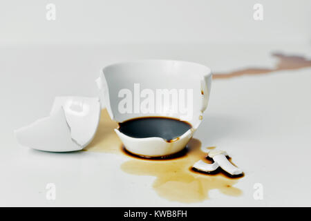a broken white ceramic cup of coffee with its pieces and the coffee spilled on a white table Stock Photo