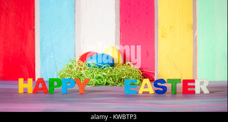 Easter banner with the words happy easter written with multicolor letters and painted eggs, on a purple table and a colorful fence in the background. Stock Photo