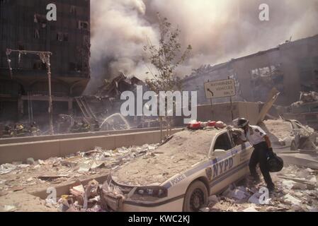 Policeman reaching into a debris covered police car after 9-11 terrorist attack in NYC. At left is still standing WTC 6. At right is the destroyed North pedestrian bridge over West Side Highway (West St.). In the background is the burning pile of the collapsed WTC 1 (North Tower). New York City, Sept. 11, 2001. (BSLOC 2015 2 52) Stock Photo