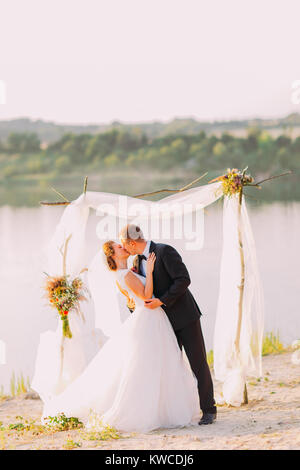 The happpy kissing newlyweds during the wedding ceremony near the river. Stock Photo