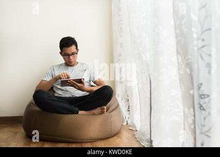 asian teenage boy using a digital tablet while relaxing on a bean bag Stock Photo