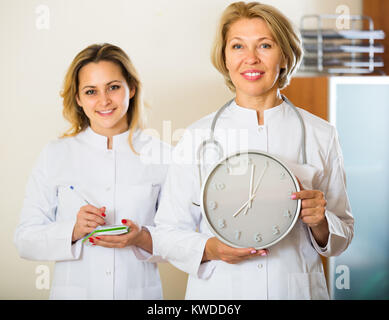 Two positive doctors in white overalls holding clock and smiling in modern clinic . Selective focus Stock Photo