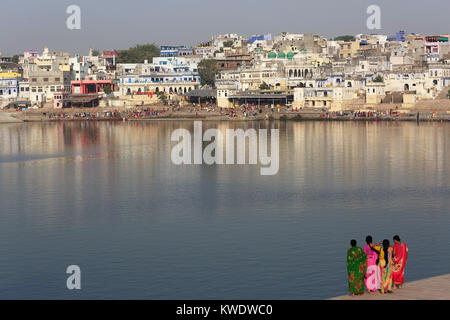 Four indian women in sari standing on the banks of holy lake at Pushkar with Hindu pilgrims taking ritual bath on the opposite side, Rajasthan, India. Stock Photo