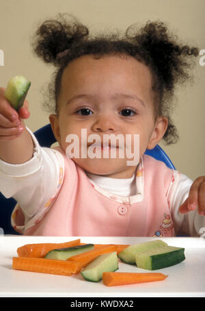 mixed race baby in high chair eating finger foods of carrot and cucumber Stock Photo