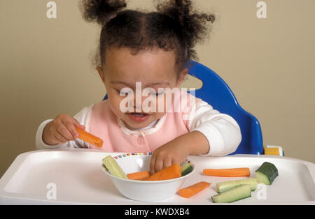 mixed race baby in high chair eating finger foods of carrot and cucumber Stock Photo