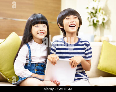 two cute asian children little boy and little girl sitting on couch holding digital tablet laughing. Stock Photo
