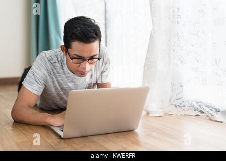 asian teen using laptop while lying on a floor in his home. weekend freelancer concept. Stock Photo