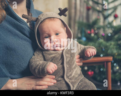 A sad baby wearing a reindeer outfit in front of the Christmas tree Stock Photo