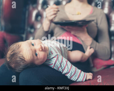 A young mother is sitting on a sofa and is trying to eat her dinner with her baby on her lap Stock Photo