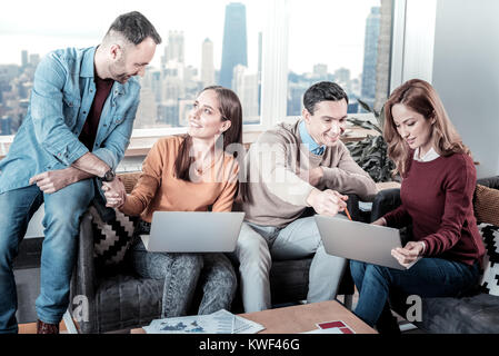 Reliable colleagues. Two nice-looking satisfied clever workers sitting in the office near their colleagues shaking their hands and using the laptop. Stock Photo