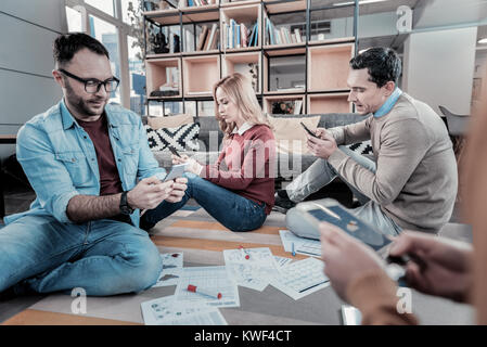 Serious and occupied. Smart concentrated busy employees sitting on the floor around papers using their smartphones and messaging. Stock Photo