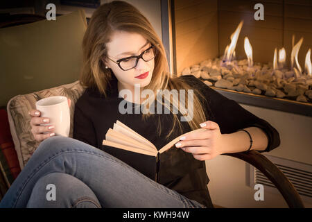 Portrait of a blonde young woman wearing eyeglasses, reading a book, sitting on a rocking chair beside a fireplace and having coffee. Stock Photo