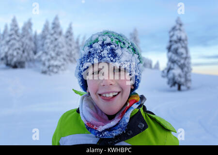 Portrait of a smiling young boy with his face and beanie covered with snow after winter playing on a mountain with spruces, Krvavec, Slovenia. Stock Photo