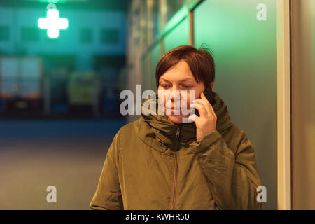 Worried woman talking on mobile phone in front of drugstore at night. Adult female using telephone to consult with doctor before buying medication in  Stock Photo