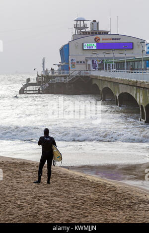 Bournemouth, Dorset, England UK. 4th January, 2018. UK weather:  surfer standing on the beach near the pier looking out to sea as surfers enjoy the surf on a windy day at Bournemouth beach, as the surfers make the most of the windy conditions and big waves. Stock Photo