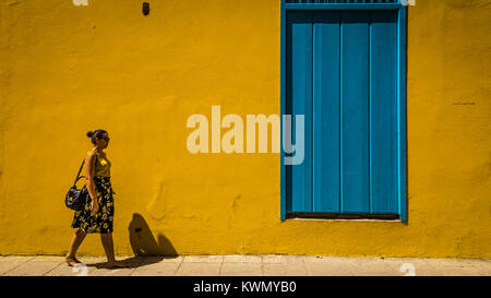 Woman walking past a colorful yellow building with bright blue closed wooden door or window in summer sunshine on a paved sidewalk Stock Photo