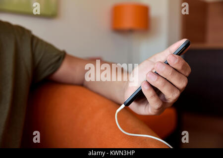 closeup of a young caucasian using a smpartphone with a cable plugged into it, sitting in a comfortable sofa indoors Stock Photo