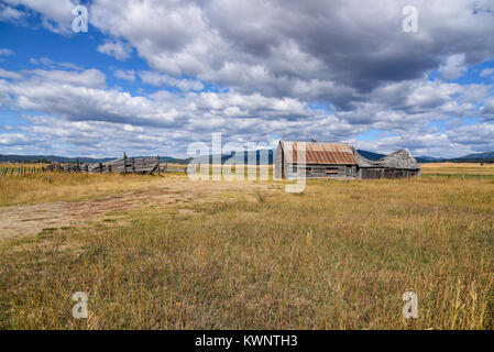 Abandoned wooden barn and farm buildings in a prairie field behind a fence next in Idaho state, USA on a sunny cloudy day Stock Photo