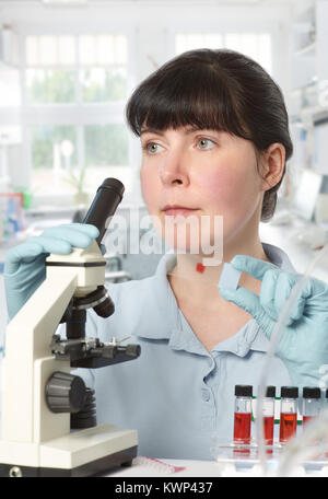 Portrait of a young female scientist analyzing red liquid samples under the microscope Stock Photo