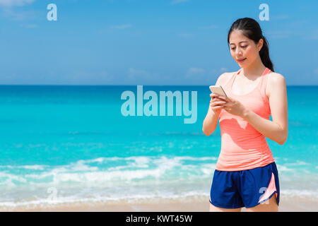 closeup photo of smiling happy female runner travel on beach going to outdoor workout and using mobile smartphone chatting with friend. Stock Photo
