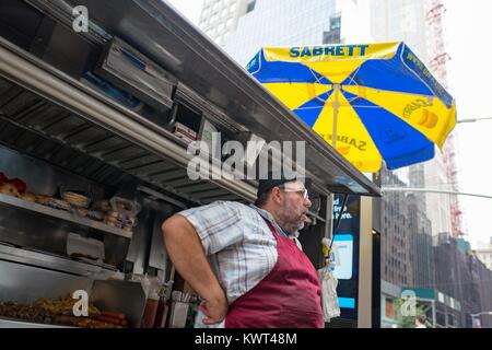 A street food vendor wearing an apron stands with his cart on 60th Street in Manhattan, New York City, New York, with hot dogs and other foods visible in the cart and a blue and yellow Sabrett umbrella visible above, September 14, 2017. Stock Photo