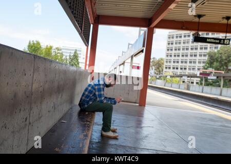 A man wearing a flannel shirt sits alone on the platform of the Walnut Creek, California station of the Bay Area Rapid Transit (BART) light rail system and uses a mobile device while waiting for a train traveling towards San Francisco, California, September 13, 2017. Stock Photo