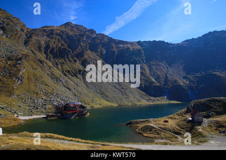 Autumn scenery landscape with water reflecting the sun rays and cabin and Fagaras Mountains in the background at Balea Lake, Sibiu County, Romania. Stock Photo