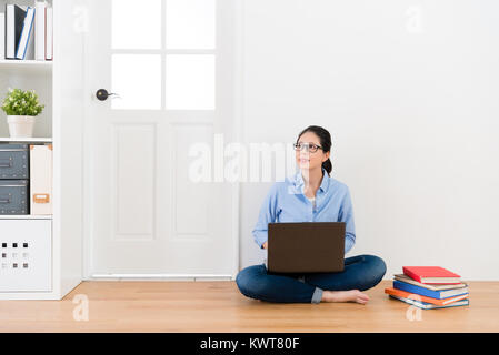 smiling beauty girl student sitting on wooden floor with white wall background and looking at air daydreaming when she using mobile computer study. Stock Photo