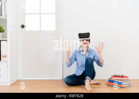 beautiful young teacher woman finished work and sitting on wooden floor wearing virtual reality goggles viewing 3D simulation video feeling surprised. Stock Photo