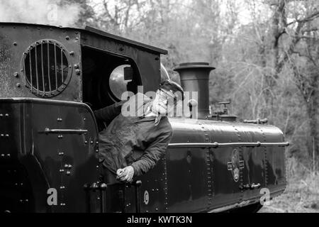 Black & white, landscape shot of moving steam train on railway line. Stood on footplate, train driver leans out of loco, checking behind. All aboard! Stock Photo