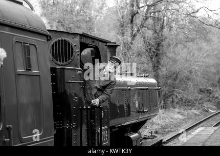 Black & white close up of vintage UK steam locomotive 7714 on railway line with engine driver on footplate leaning out of train. All aboard! Stock Photo