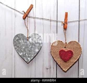 Two hearts, one silver and the other one brown with a smaller red heart in it's center, hanging on a line in front of a white washed fence Stock Photo