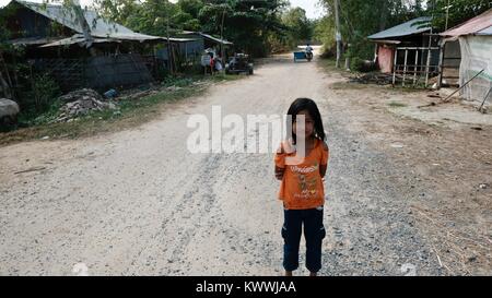 Rural Takeo Cambodia Mekong Delta little girl with long black hair stranding in the middle of a dirt road wearing an orange tee shirt and black pants Stock Photo