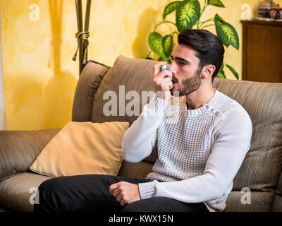 Handsome young man using inhaler for asthma, while sitting on sofa at home. Stock Photo