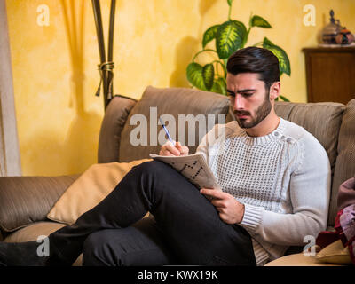 Young man sitting doing a crossword puzzle looking thoughtfully at a magazine, with his pencil to his mouth, as he tries to think of the answer to the Stock Photo