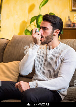 Handsome young man using inhaler for asthma, while sitting on sofa at home. Stock Photo