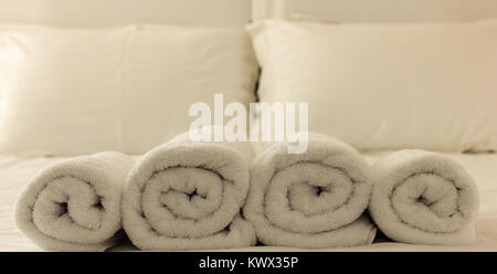 Hotel bedroom. White fluffy, rolled towels in row, linen beddings on tidy bed. Close up view. Stock Photo