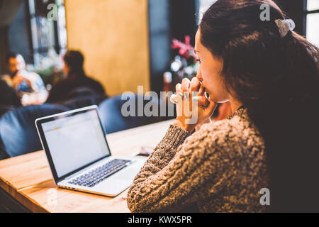 beautiful young brunette girl in sweater working on laptop computer inside cafe at wooden table near window. Winter and Christmas decorations. The con Stock Photo