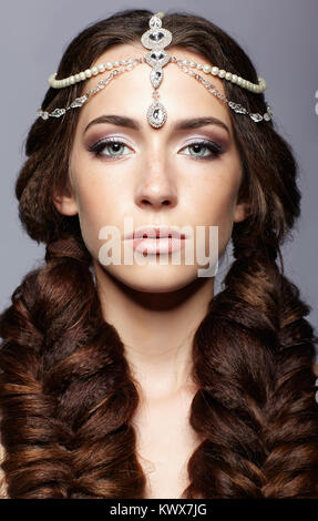 Beauty portrait of young woman with diadem. Brunette girl with long hair plaits and day female makeup on gray background. Stock Photo