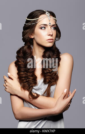 Beauty portrait of young woman with diadem. Brunette girl with long hair plaits and day female makeup on gray background. Stock Photo