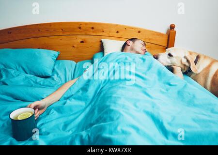 Influenza season. Sick man under blanket is holding cup of the hot tea and looking on his dog. Stock Photo