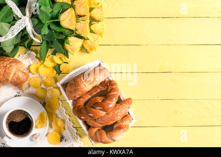 Breakfast table with a cup of coffee, freshly baked croissants and bagels, with flower petals on the yellow wooden table and a big bouquet of roses. Stock Photo