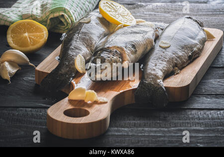 Close-up image with fresh trout displayed on a wooden cutting board, seasoned with garlic and lemon slices, on a rustic wooden table, on a sunny day. Stock Photo